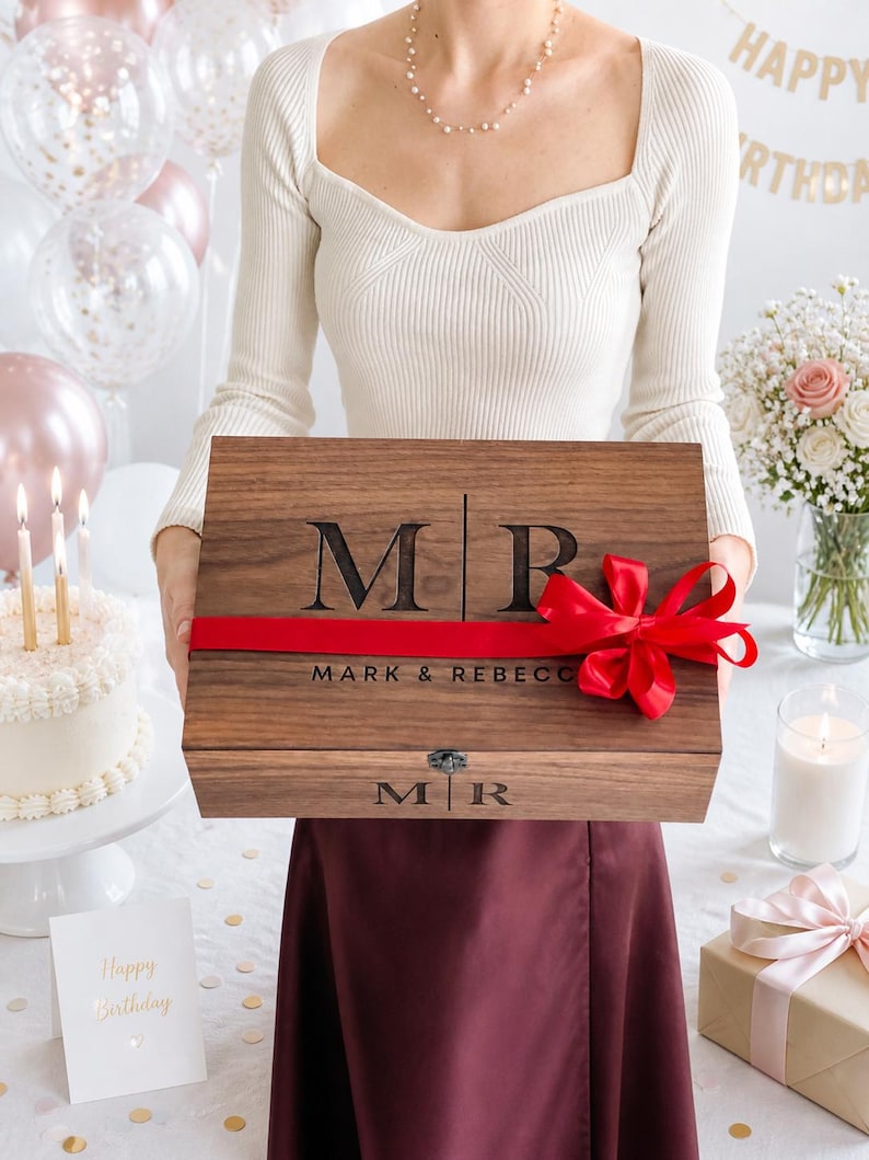 May include: A wooden gift box with the initials "MR" and "MARK & REBECCA" engraved on the lid. The box is tied with a red ribbon and bow. The background includes a birthday cake, balloons, and a "Happy Birthday" card.