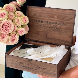 May include: A wooden keepsake box with the inscription "OUR HAPPILY ever after" on the lid. Inside are white feathers and envelopes. The box is engraved with "JOSEPH + KAYLA" and the date "30 | JULY | 2023". A bouquet of pink roses is held nearby.