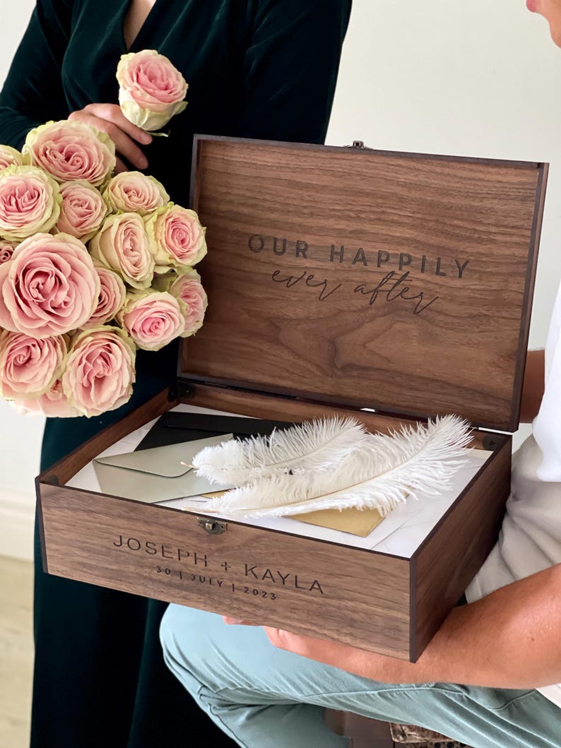 May include: A wooden keepsake box with the inscription "OUR HAPPILY ever after" on the lid. Inside are white feathers and envelopes. The box is engraved with "JOSEPH + KAYLA" and the date "30 | JULY | 2023". A bouquet of pink roses is held nearby.