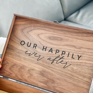 May include: A wooden box with the words "OUR HAPPILY ever after" printed on the inside of the lid. The box is made of a dark wood with a visible grain pattern. The text is in a black, elegant font.