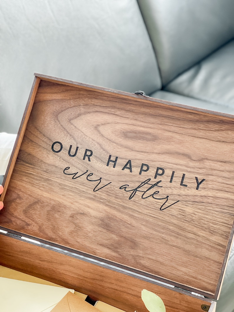 May include: A wooden box with the words "OUR HAPPILY ever after" printed on the inside of the lid. The box is made of a dark wood with a visible grain pattern. The text is in a black, elegant font.