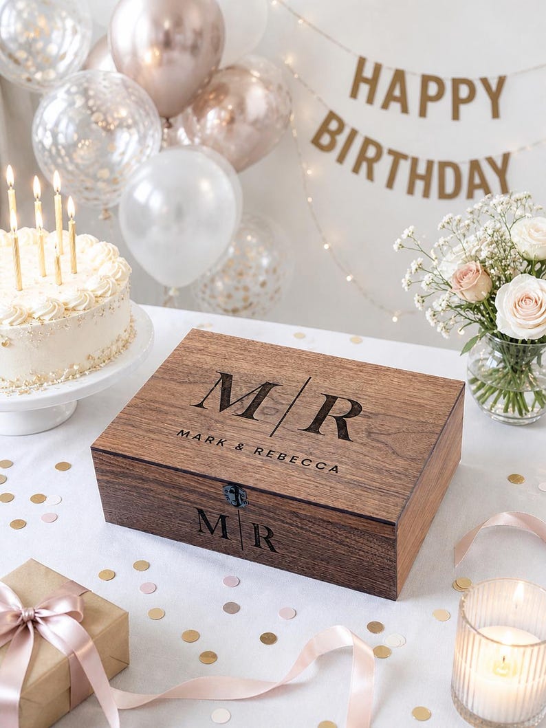 May include: A wooden box with the initials "MR" and the names "Mark & Rebecca" engraved on the lid. The box is on a white surface with confetti, a gift box, a lit candle, and a birthday cake. Balloons and a "Happy Birthday" banner are in the background.
