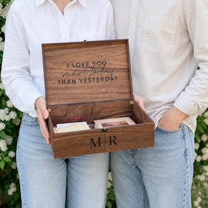 May include: A wooden keepsake box with the inscription "I love you more today than yesterday" engraved on the inside lid. The box is open, revealing contents and the initials "MR" on the front. The box is held by two people.