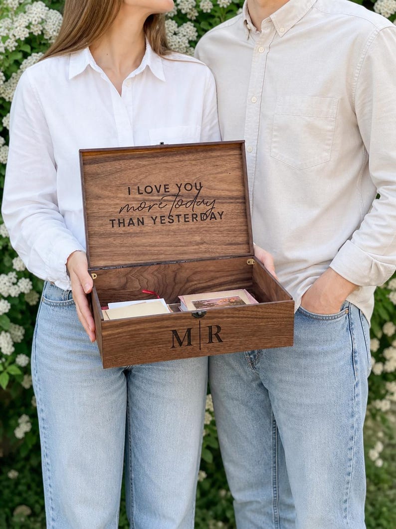 May include: A wooden keepsake box with the inscription "I love you more today than yesterday" engraved on the inside lid. The box is open, revealing contents and the initials "MR" on the front. The box is held by two people.