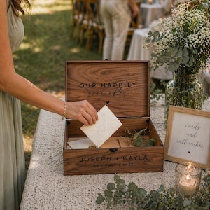 May include: A wooden wedding card box with the words "OUR HAPPILY ever after" engraved on the lid. The box is open, revealing cards inside. The names "JOSEPH + KAYLA" and the date "30 JULY 2023" are also engraved on the box.
