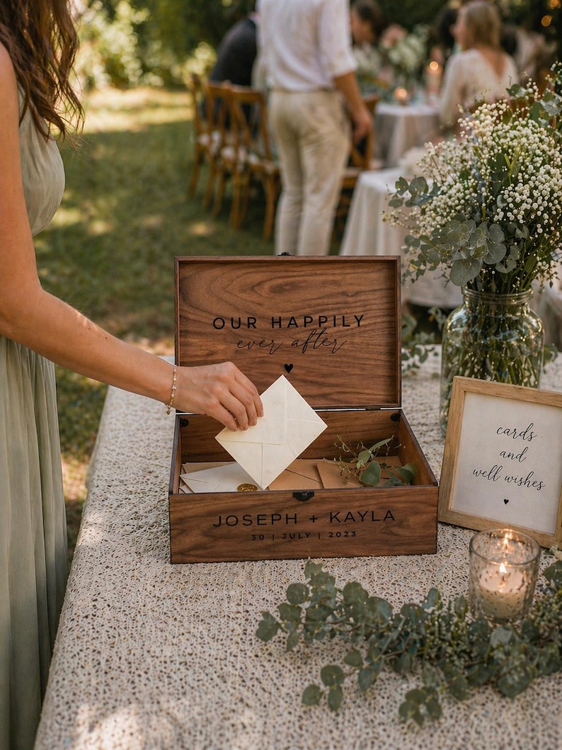 May include: A wooden wedding card box with the words "OUR HAPPILY ever after" engraved on the lid. The box is open, revealing cards inside. The names "JOSEPH + KAYLA" and the date "30 JULY 2023" are also engraved on the box.