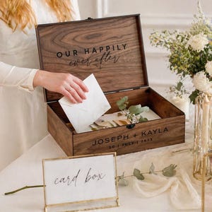 May include: A wooden card box with the words "OUR HAPPILY ever after" engraved on the lid. The box is open, revealing cards and greenery inside. The names "JOSEPH + KAYLA" and the date "30 | JULY | 2023" are also engraved. A sign that says "card box" is in front.