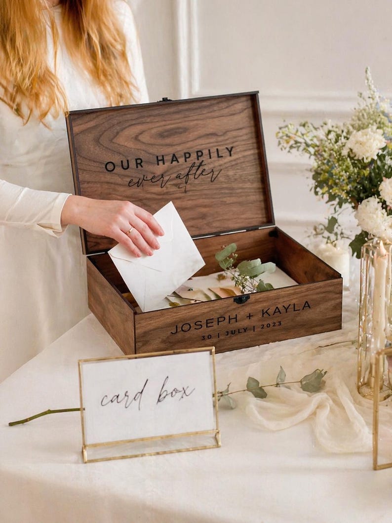 May include: A wooden card box with the words "OUR HAPPILY ever after" engraved on the lid. The box is open, revealing cards and greenery inside. The names "JOSEPH + KAYLA" and the date "30 | JULY | 2023" are also engraved. A sign that says "card box" is in front.