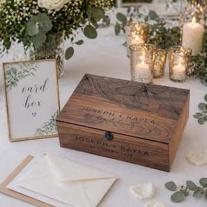 May include: A wooden card box with floral design and the names "JOSEPH + KAYLA" engraved on it. The box is on a table with a "card box" sign, envelopes, and candles. The date "30 | JULY | 2023" is also engraved.