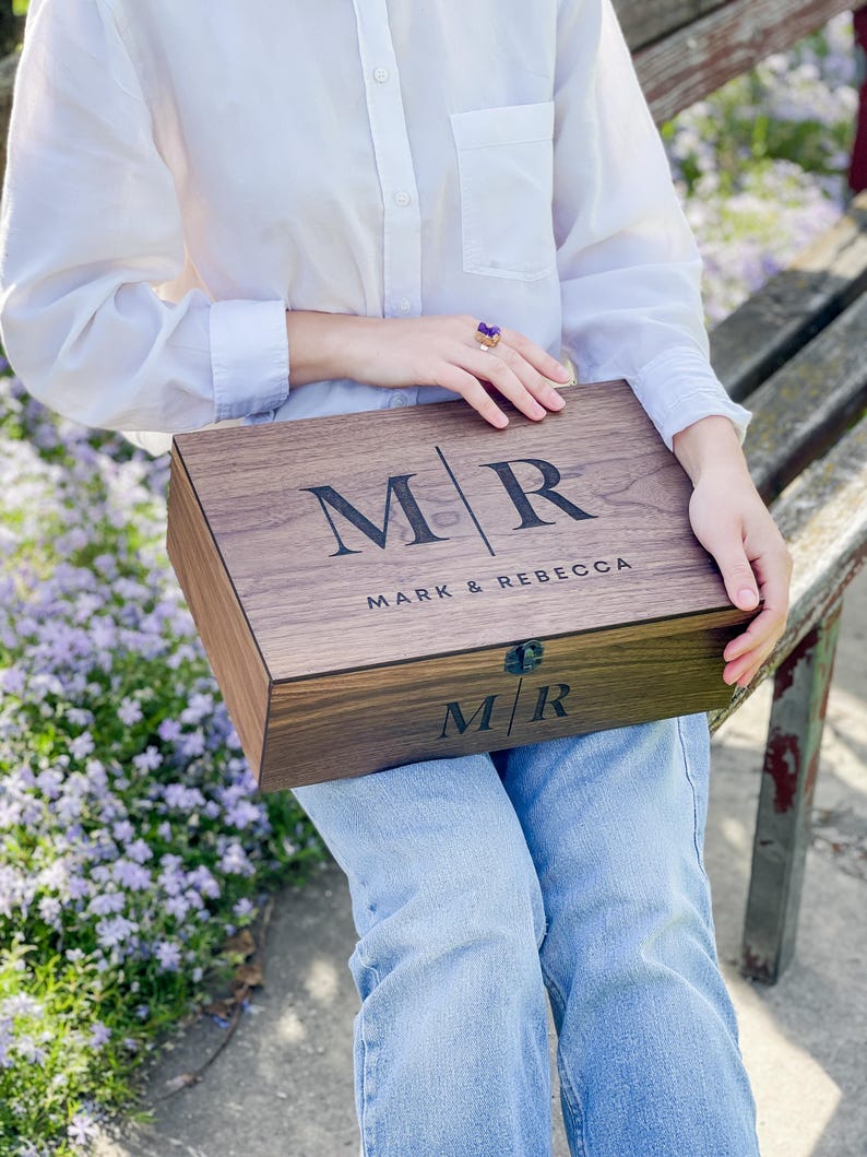 May include: A wooden box with the initials "MR" and the names "MARK & REBECCA" engraved on the lid. The box is a dark brown color and has a rectangular shape. The box is being held by someone wearing a white shirt and blue jeans.