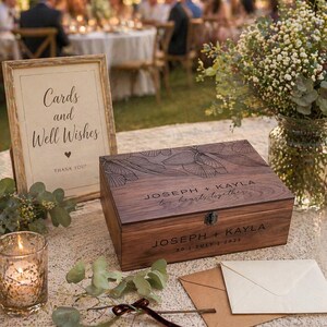 May include: A wooden keepsake box with engraved floral design and the names "JOSEPH + KAYLA" and the date "30 | JULY | 2023". A sign reads "Cards and Well Wishes". A lit candle and greenery are also present.