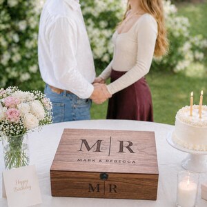 May include: A wooden box with the initials "MR" and the names "Mark & Rebecca" engraved on the lid. The box is on a white tablecloth, with a bouquet of flowers and a cake with candles in the background.