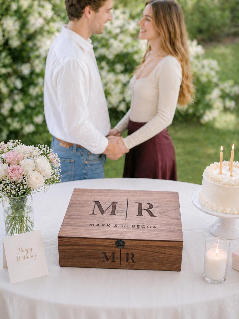 May include: A wooden box with the initials "MR" and the names "Mark & Rebecca" engraved on the lid. The box is on a white tablecloth, with a bouquet of flowers and a cake with candles in the background.