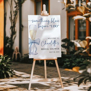 May include: A white sign with blue text and a watercolor illustration of a champagne flute with a blue ribbon. The sign reads 'Something blue before 'I DO'', 'Welcome to Charlotte's Bridal Shower', and 'May 18, 2025'.