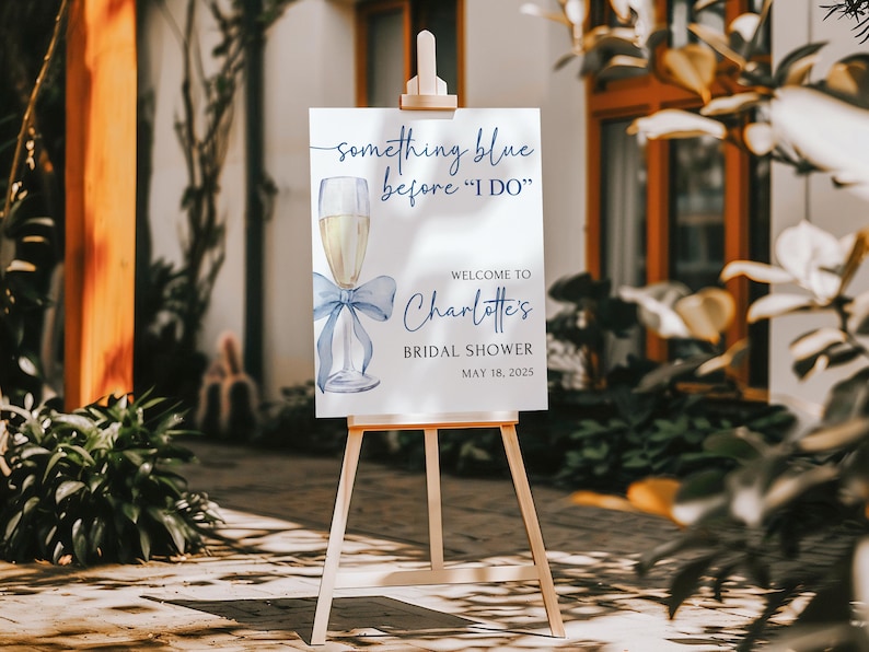 May include: A white sign with blue text and a watercolor illustration of a champagne flute with a blue ribbon. The sign reads 'Something blue before 'I DO'', 'Welcome to Charlotte's Bridal Shower', and 'May 18, 2025'.
