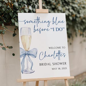 May include: A white sign with blue text and a blue watercolor illustration of a champagne flute with a blue bow. The text reads 'something blue before 'I DO'', 'WELCOME TO Charlotte's', 'BRIDAL SHOWER', and 'MAY 18, 2025'.