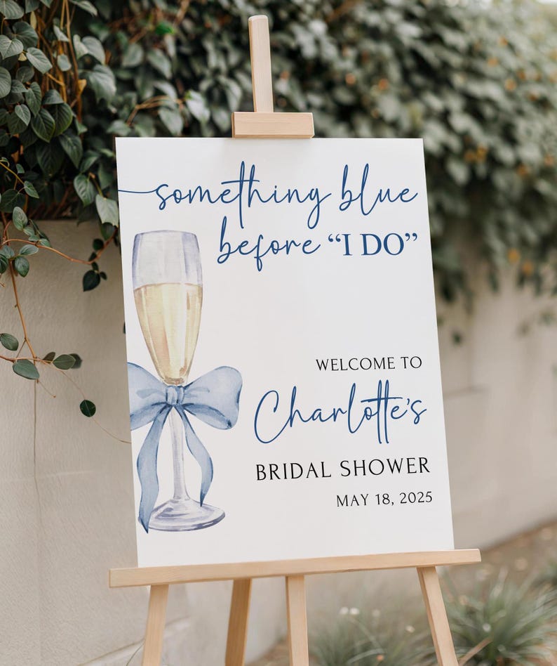 May include: A white sign with blue text and a blue watercolor illustration of a champagne flute with a blue bow. The text reads 'something blue before 'I DO'', 'WELCOME TO Charlotte's', 'BRIDAL SHOWER', and 'MAY 18, 2025'.