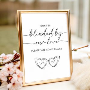 May include: A framed sign with a gold border displays the text "DON'T BE blinded by our love" and "PLEASE TAKE SOME SHADES." Below the text is a drawing of heart-shaped sunglasses. The sign is on a white surface with flowers.