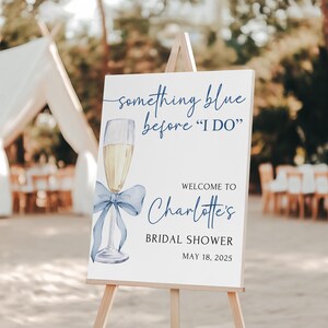 May include: A white sign with blue text and a blue watercolor illustration of a champagne flute with a blue bow. The sign reads 'something blue before 'I DO'', 'Welcome to Charlotte's Bridal Shower', and 'May 18, 2025'.
