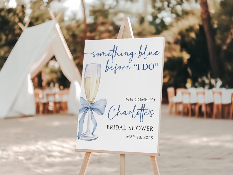 May include: A white sign with blue text and a blue watercolor illustration of a champagne flute with a blue bow. The sign reads 'something blue before 'I DO'', 'Welcome to Charlotte's Bridal Shower', and 'May 18, 2025'.
