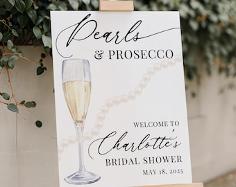 May include: A white sign with black text and a watercolour illustration of a champagne flute and pearls. The text reads "Pearls & Prosecco" and "Welcome to Charlotte's Bridal Shower May 18, 2025".