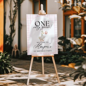 Puede incluir: Un caballete de madera exhibe un letrero con el texto "ONE silly goose!" encima de un dibujo de un ganso sosteniendo un globo. El letrero da la bienvenida a "Harper's 1st Birthday Party" en un marco a cuadros rosa y blanco.