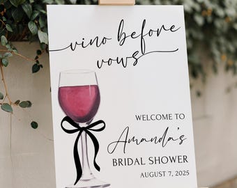 May include: A white sign on a wooden easel with the text "vino before vows" and a wine glass illustration. The sign welcomes guests to Amanda's bridal shower on August 7, 2025. A black bow is tied around the wine glass.