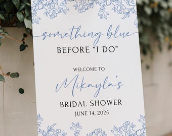 May include: A white sign on a wooden easel features blue floral designs and text. The sign says "something blue BEFORE 'I DO' WELCOME TO Mikayla's BRIDAL SHOWER JUNE 14, 2025". A decorative element for a bridal shower.