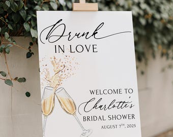 May include: A white sign on a wooden easel, featuring the text "Drunk in Love" in cursive. It also reads "Welcome to Charlotte's Bridal Shower, August 7th, 2025." Two champagne glasses are illustrated with gold details. The sign is displayed against a backdrop of foliage.