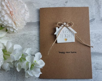 May include: A brown card with a white house-shaped ornament attached with twine. The ornament says "NEW HOME" and has a small gold heart. The card says "happy new home" in black ink.