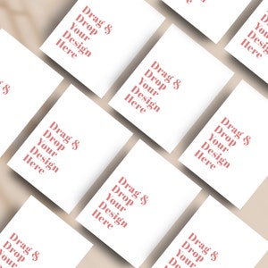 May include: A collection of white rectangular cards arranged diagonally on a light beige surface. Each card features the text "Drag & Drop Your Design Here" in a soft red hue, creating a visually appealing layout.