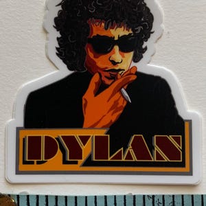 May include: A sticker featuring a colourful illustration of Bob Dylan with the word "DYLAN" in a stylised font below. The sticker is approximately 5 cm wide.