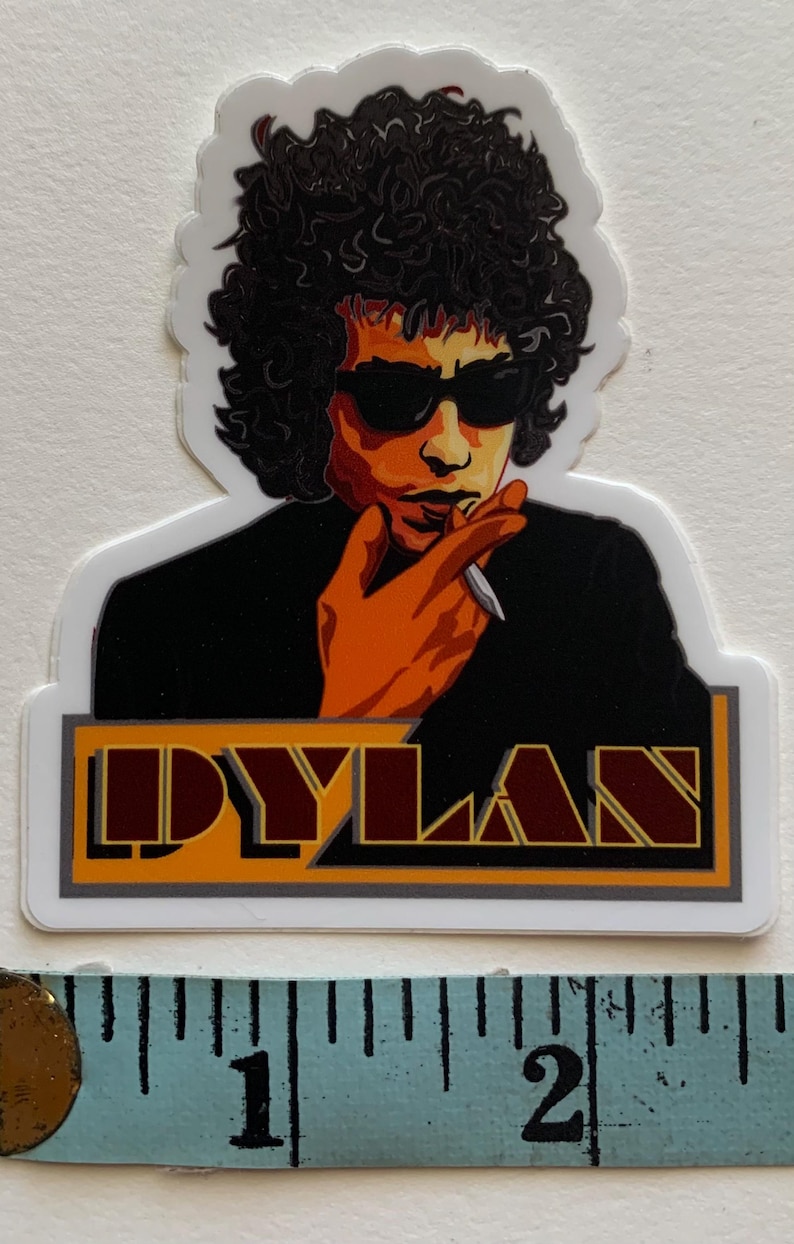 May include: A sticker featuring a colourful illustration of Bob Dylan with the word "DYLAN" in a stylised font below. The sticker is approximately 5 cm wide.