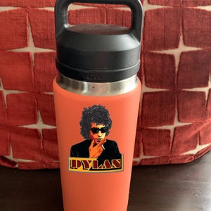 May include: A coral-coloured Yeti water bottle with a black lid and handle. The bottle has a sticker of Bob Dylan with the text "DYLAN" in a gold and red banner.