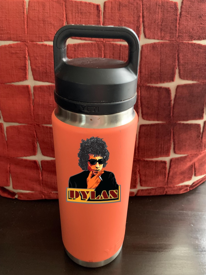 May include: A coral-coloured Yeti water bottle with a black lid and handle. The bottle has a sticker of Bob Dylan with the text "DYLAN" in a gold and red banner.