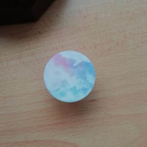 Pode incluir: Um suporte de telefone redondo com um design de nuvem de aquarela azul e rosa.