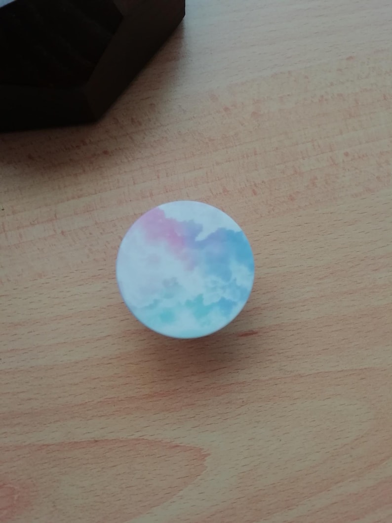 Pode incluir: Um suporte de telefone redondo com um design de nuvem de aquarela azul e rosa.