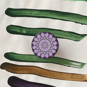 Pode incluir: Um design de mandala roxo e branco sobre um fundo preto. O mandala &eacute; cercado por quatro pinceladas verdes sobre um fundo branco.