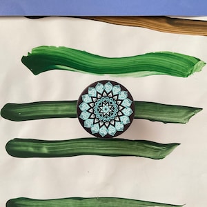 Pode incluir: Um agarre de telefone mandala preto e branco com um design azul e branco. O agarre est&aacute; sobre uma superf&iacute;cie branca com pinceladas verdes.