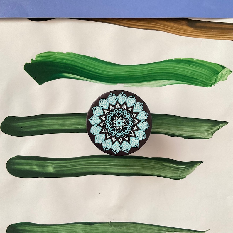 Pode incluir: Um agarre de telefone mandala preto e branco com um design azul e branco. O agarre est&aacute; sobre uma superf&iacute;cie branca com pinceladas verdes.