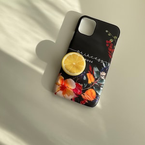 Pode incluir: Capa de telefone preta com um design floral com flores vermelhas, laranja, amarelas e azuis. O texto "princess rose" est&aacute; impresso na capa. Uma fatia de lim&atilde;o est&aacute; em cima da capa.