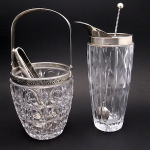 May include: A clear, cut-glass ice bucket with silver-tone handle and tongs, next to a matching cocktail shaker with a silver-tone rim and bar spoon. The set is on a black background.