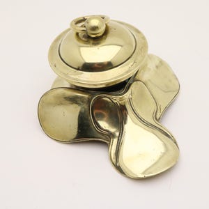 Pode incluir: Um tinteiro de latão vintage com uma tampa em cúpula e uma pequena pega em anel. O tinteiro tem uma base única de três lóbulos. O latão tem um acabamento polido, refletindo a luz. O design é ornamentado e provavelmente de uma época anterior.