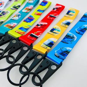 K&ouml;nnte beinhalten: Ein Set bunter Schl&uuml;sselanh&auml;nger mit verschiedenen Rennhelm-Designs. Jeder Schl&uuml;sselanh&auml;nger hat eine andere Farbe und ein anderes Helmdesign, darunter Blau, Gelb, Rot, Gr&uuml;n und Wei&szlig;. Die Schl&uuml;sselanh&auml;nger haben schwarze Metall-Drehclips und -ringe.