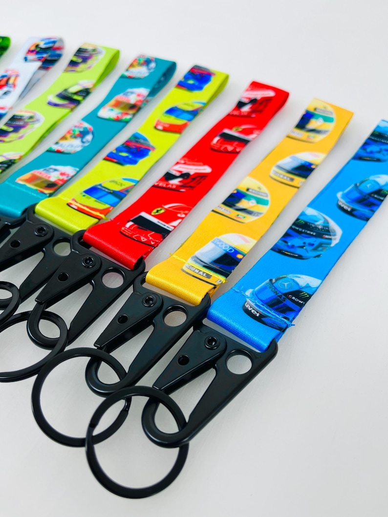 K&ouml;nnte beinhalten: Ein Set bunter Schl&uuml;sselanh&auml;nger mit verschiedenen Rennhelm-Designs. Jeder Schl&uuml;sselanh&auml;nger hat eine andere Farbe und ein anderes Helmdesign, darunter Blau, Gelb, Rot, Gr&uuml;n und Wei&szlig;. Die Schl&uuml;sselanh&auml;nger haben schwarze Metall-Drehclips und -ringe.