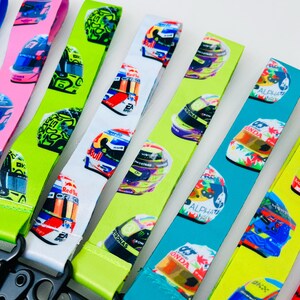 K&ouml;nnte beinhalten: Ein Set bunter Lanyards mit verschiedenen Rennhelmdesigns. Die Lanyards sind aus einem weichen, strapazierf&auml;higen Material gefertigt und in verschiedenen Farben erh&auml;ltlich, darunter Gr&uuml;n, Blau, Wei&szlig; und Pink. Jede Lanyard hat einen schwarzen Metallclip.