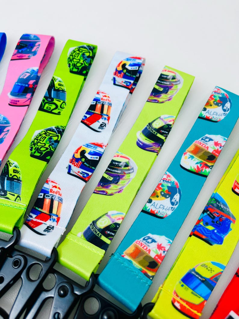 K&ouml;nnte beinhalten: Ein Set bunter Lanyards mit verschiedenen Rennhelmdesigns. Die Lanyards sind aus einem weichen, strapazierf&auml;higen Material gefertigt und in verschiedenen Farben erh&auml;ltlich, darunter Gr&uuml;n, Blau, Wei&szlig; und Pink. Jede Lanyard hat einen schwarzen Metallclip.