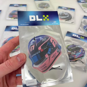 Puede incluir: Un ambientador de coche rosa y azul con un dise&ntilde;o de casco de coche de carreras. El ambientador tiene el texto "MYPROTEIN", "DURACELL", "WILLIAMS RACING" y "MONSOON VALLET". El ambientador est&aacute; etiquetado como "frambuesa".