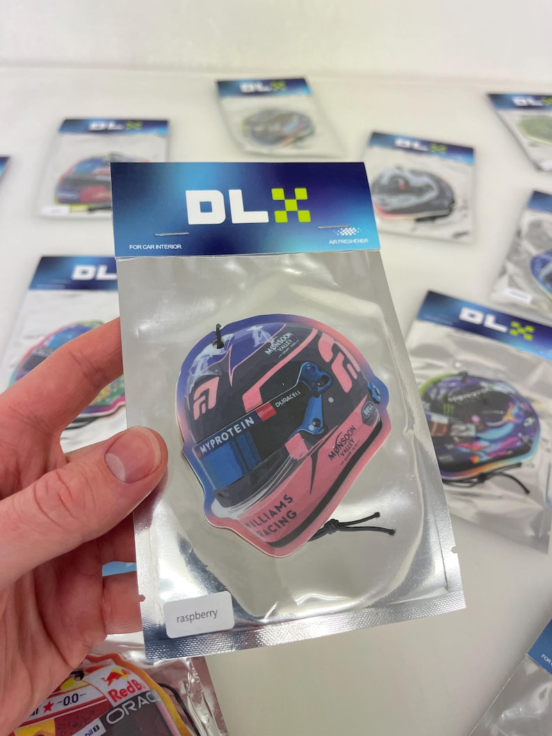 Puede incluir: Un ambientador de coche rosa y azul con un dise&ntilde;o de casco de coche de carreras. El ambientador tiene el texto "MYPROTEIN", "DURACELL", "WILLIAMS RACING" y "MONSOON VALLET". El ambientador est&aacute; etiquetado como "frambuesa".