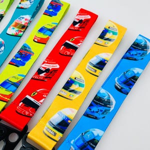K&ouml;nnte beinhalten: F&uuml;nf bunte Lanyards mit verschiedenen Designs von Rennwagenhelmen. Die Lanyards sind aus weichem, gewebtem Stoff gefertigt und haben einen schwarzen Metallclip.